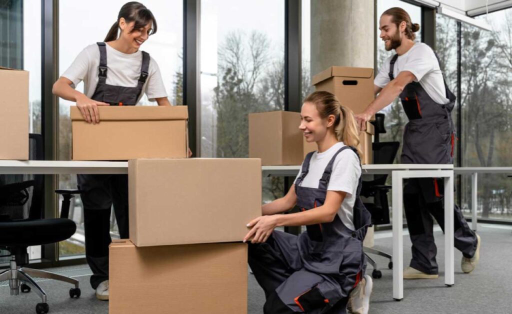 How Commercial Movers Relocate an Office Without Derailing Daily Operations Three movers in overalls packing and stacking cardboard boxes in a bright office setting.”,