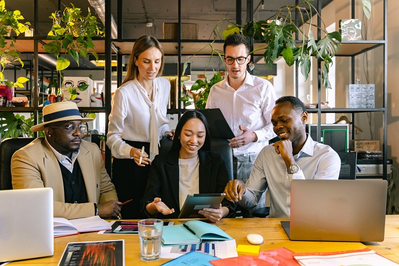 The Role of Managers in Creating More Inclusive Workplaces Five coworkers collaborate around a wooden table with laptops and tablets in a modern office filled with plants, brainstorming together.