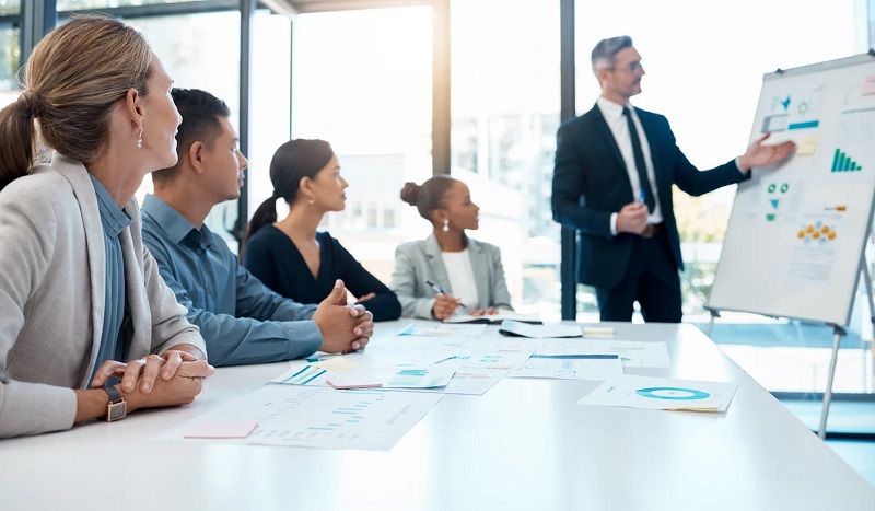 Building a Scalable Business Model: A Guide for Growing Entrepreneurs A diverse group of professionals sits around a conference table watching a presenter point to a whiteboard with charts and graphs.